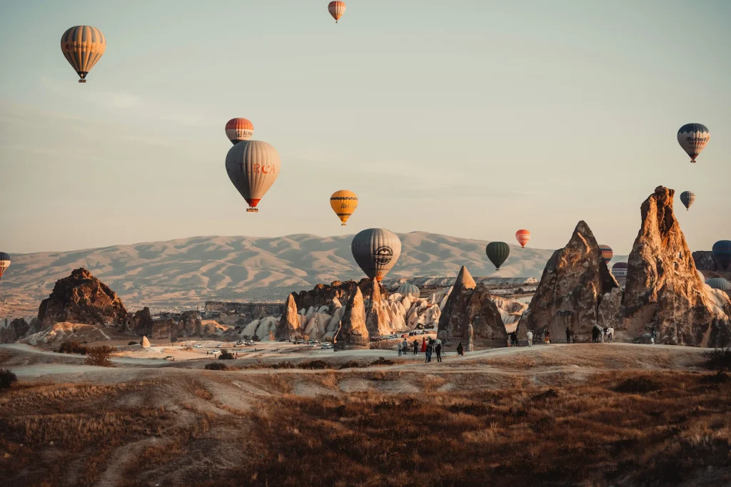 Multiple hot air balloons floating low over jagged fairy chimneys and desert valley terrain in Cappadocia during a warm golden hour sunrise.